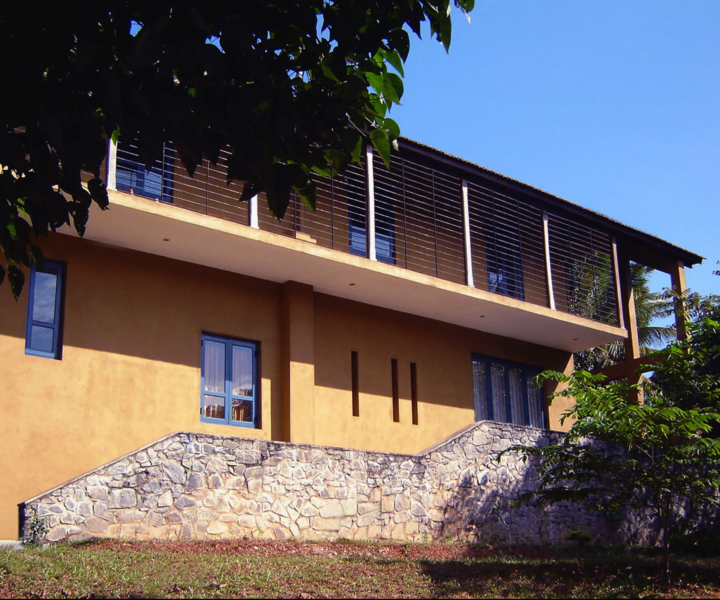 A two-storey ochre house with blue window frames and a screened upper veranda.
