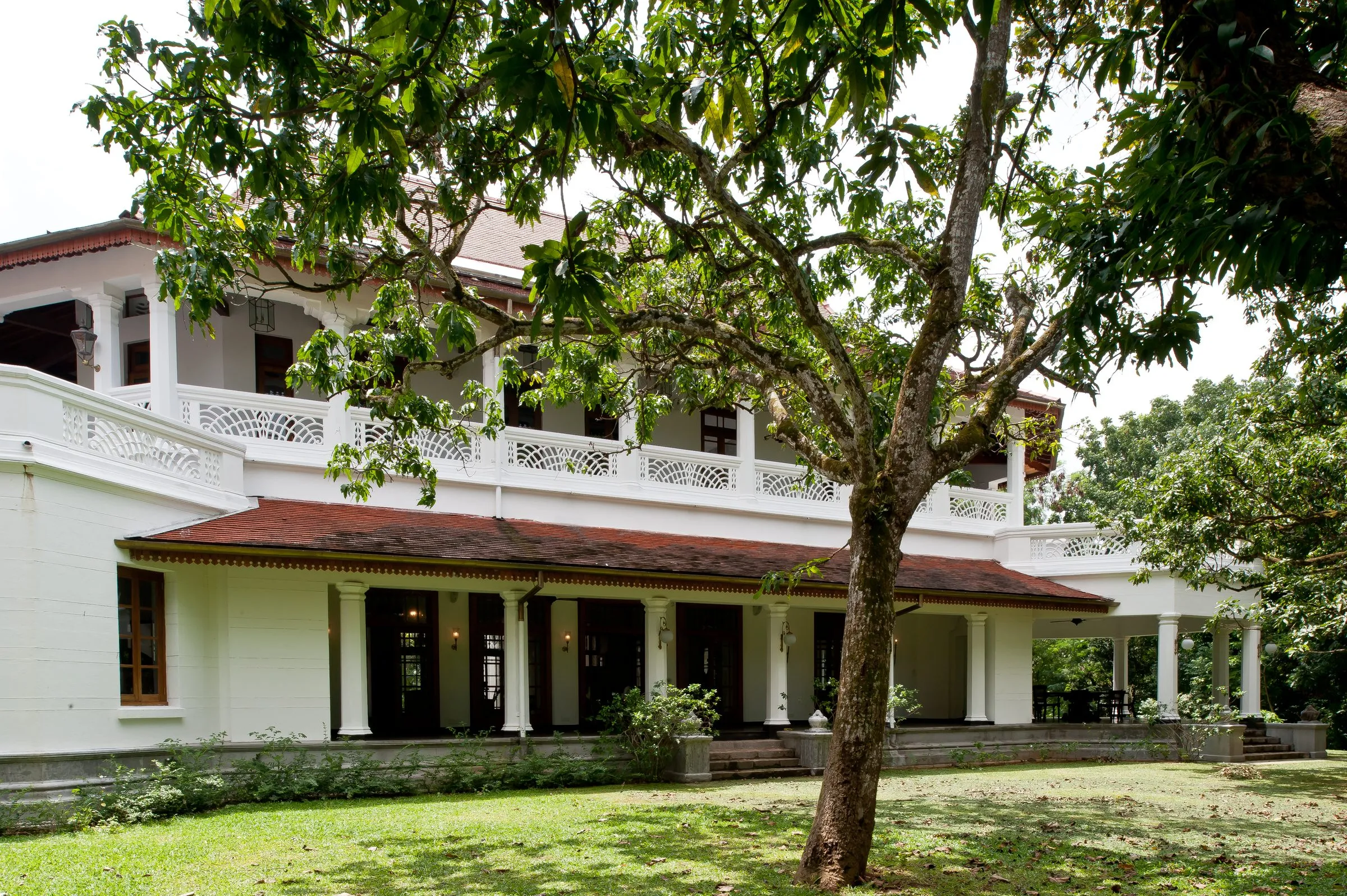 A white two-storey house with a pierced balustrade and a red-tiled veranda roof, with a large tree on the lawn.