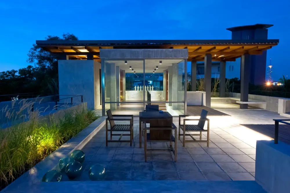 A roof terrace at night: a glazed pavilion with a dining table, planting and lit paving.
