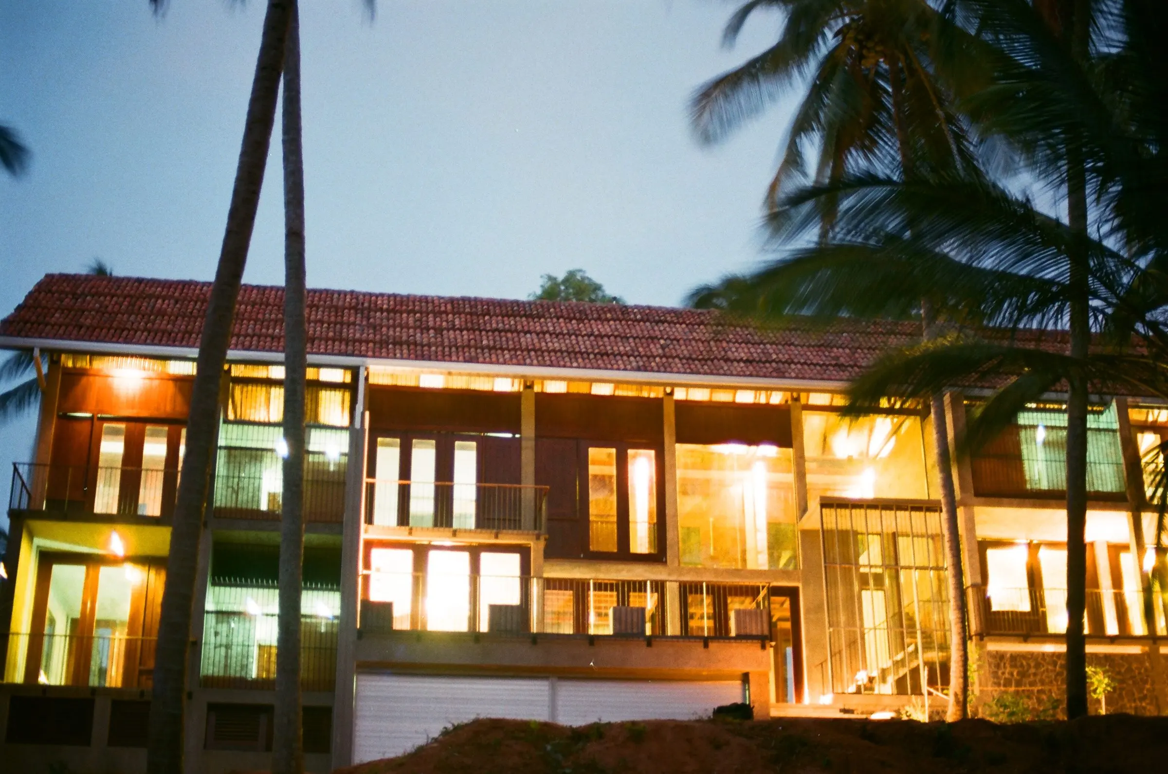 The house at dusk, with lit glazing under a tiled roof, between coconut palms.