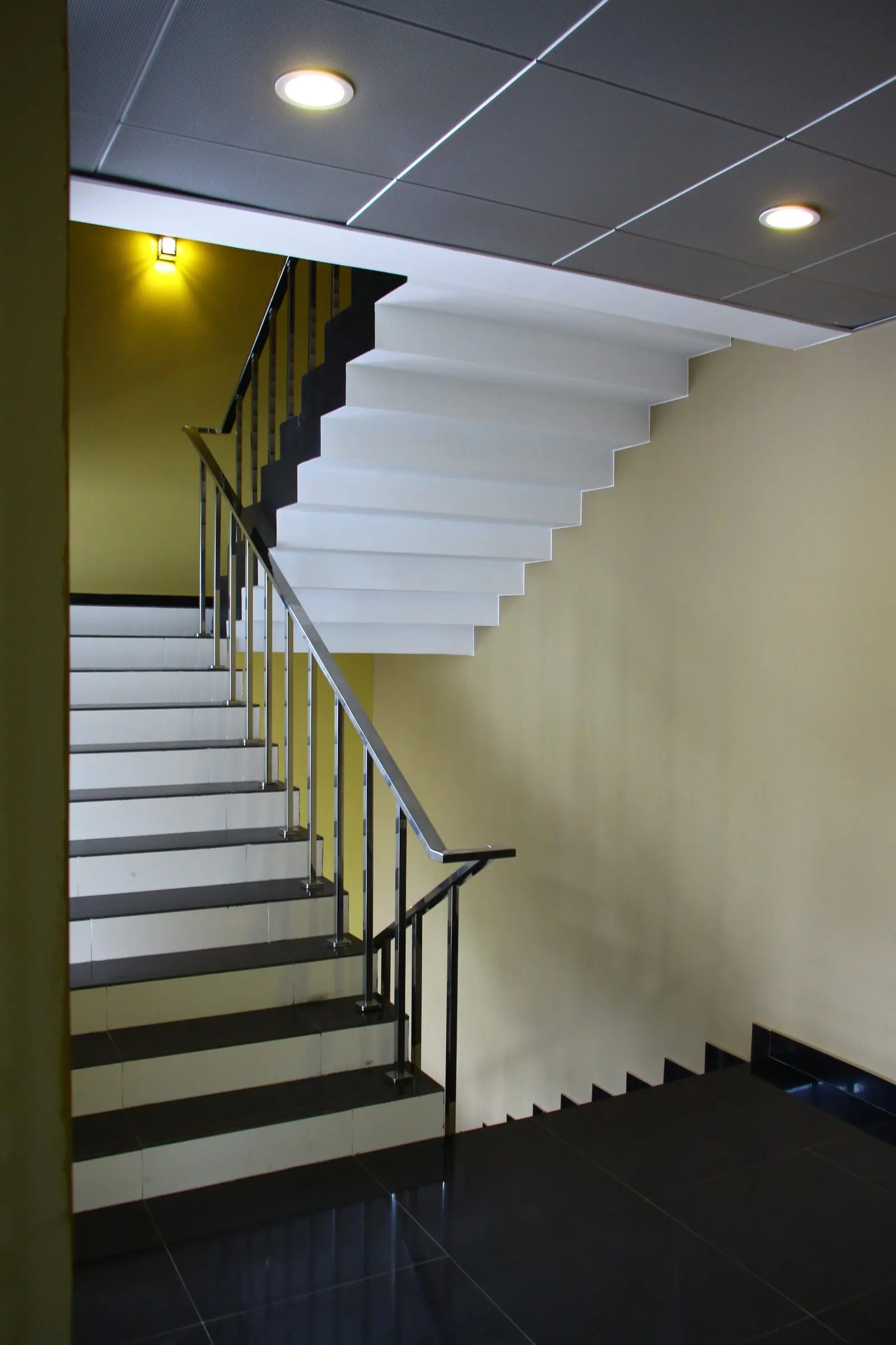 A stair hall with white treads, a black balustrade, a yellow wall and recessed ceiling lights.