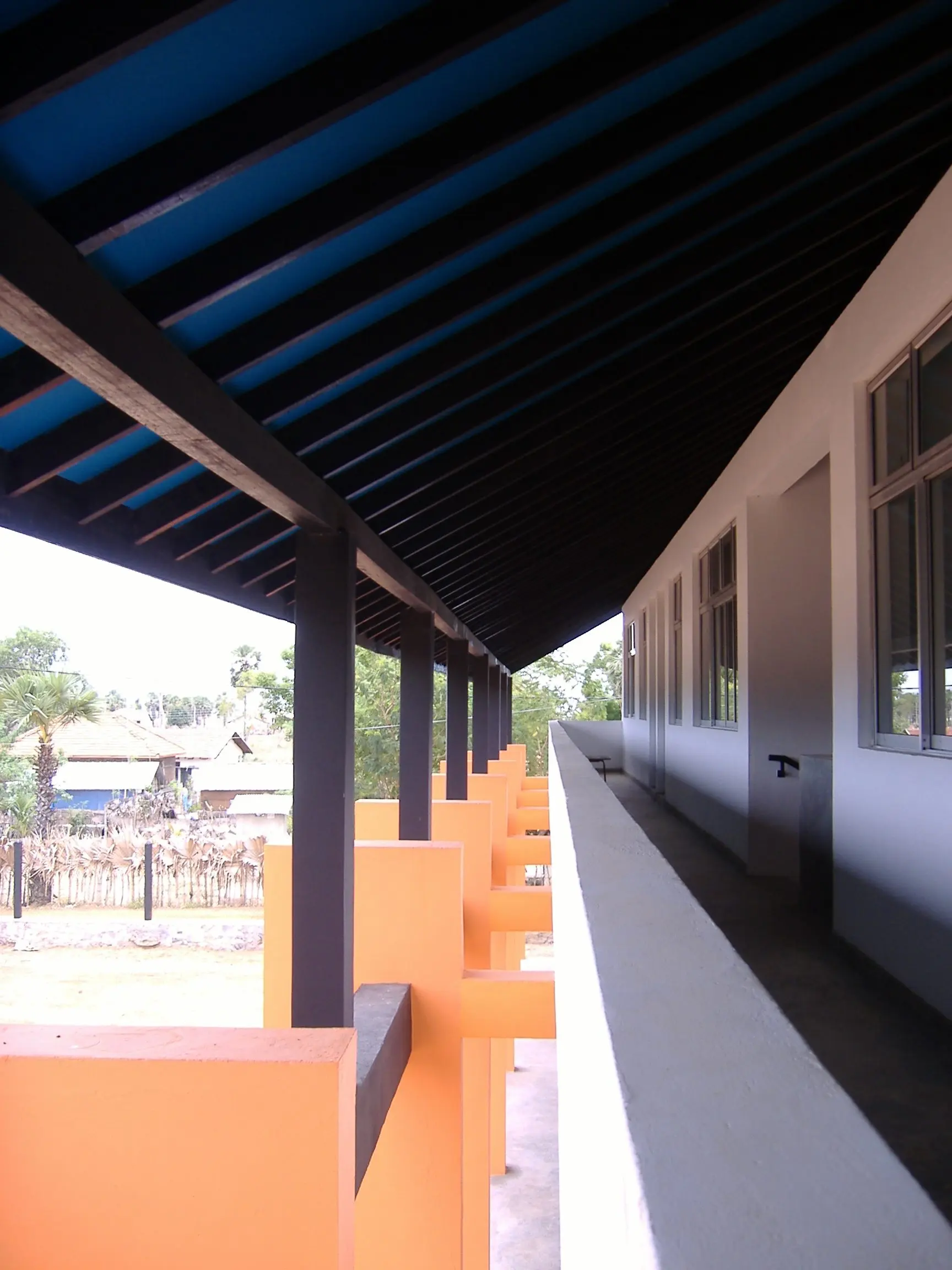 A covered walkway under a blue-lined roof, beside a white wall and orange columns.