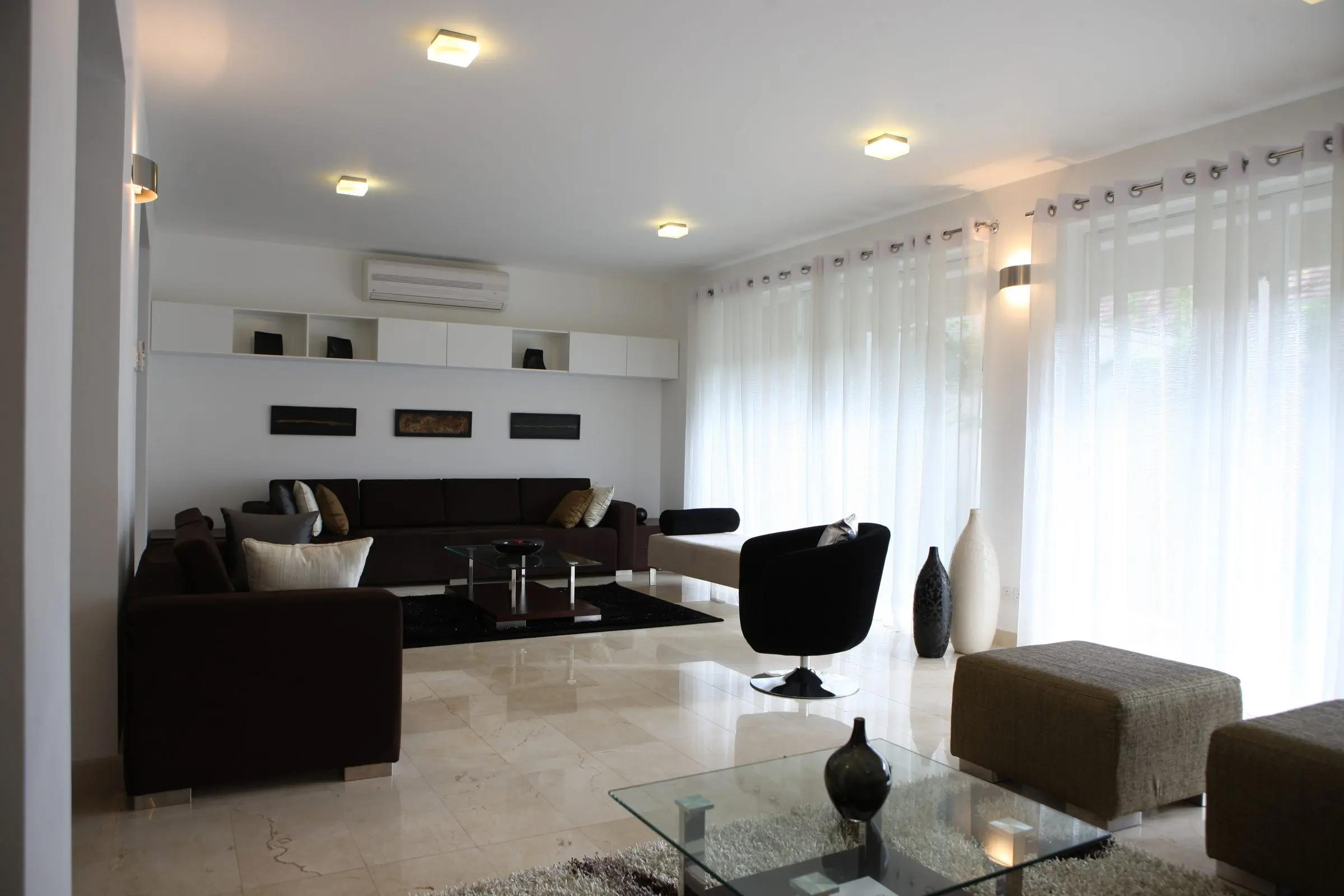 A living room with dark sofas, a coffee table and tall white curtains over full-height glazing.