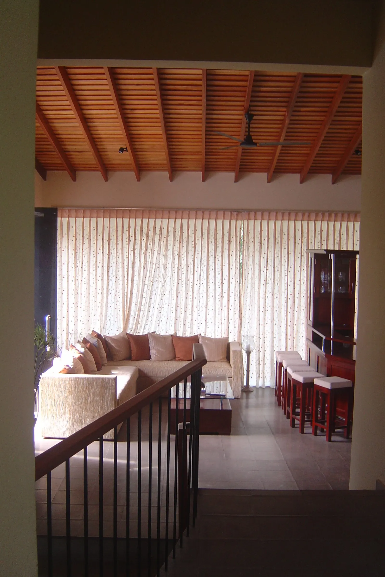 A living room under a battened ceiling, with a sectional sofa, bar stools and a stair rail in the foreground.