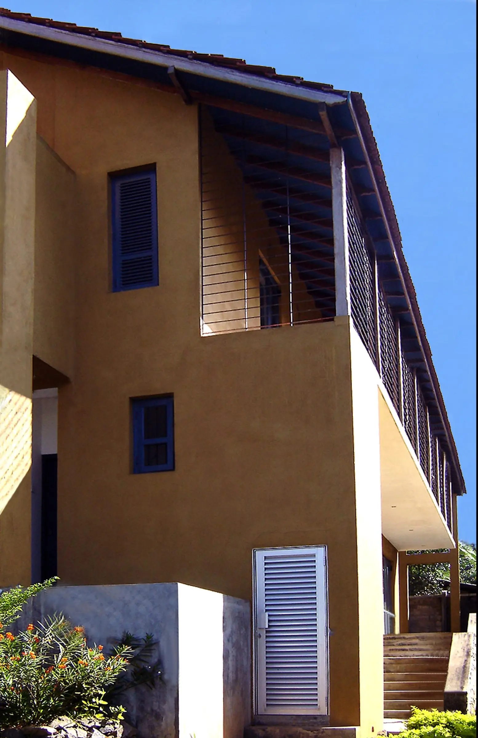 A side elevation with ochre walls, blue shutters, a tiled roof and a screened upper balcony.