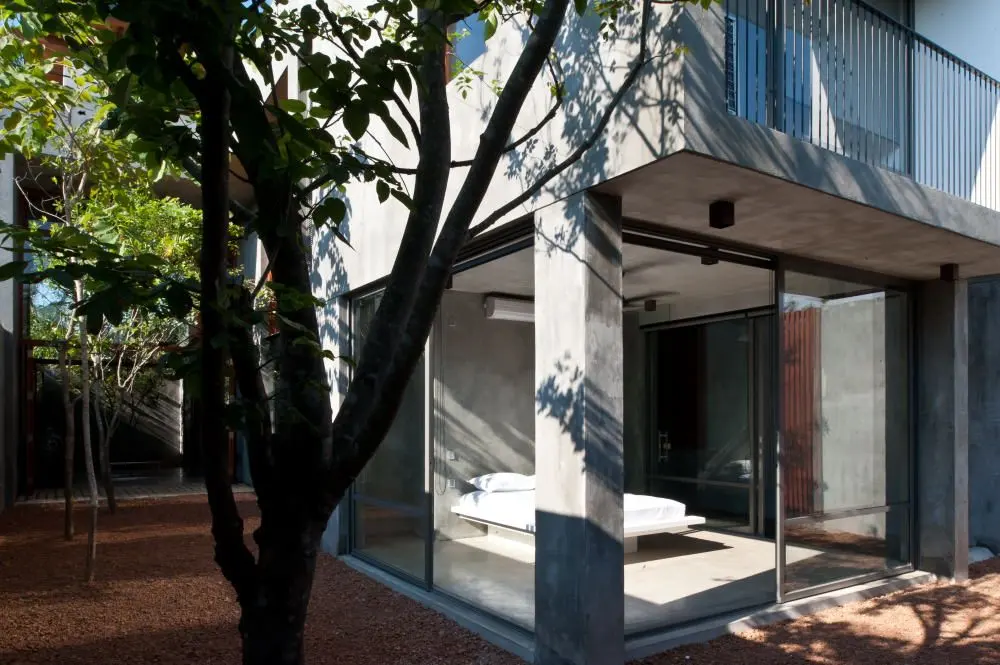 A courtyard with a tree beside a glazed bedroom, with a balcony above.