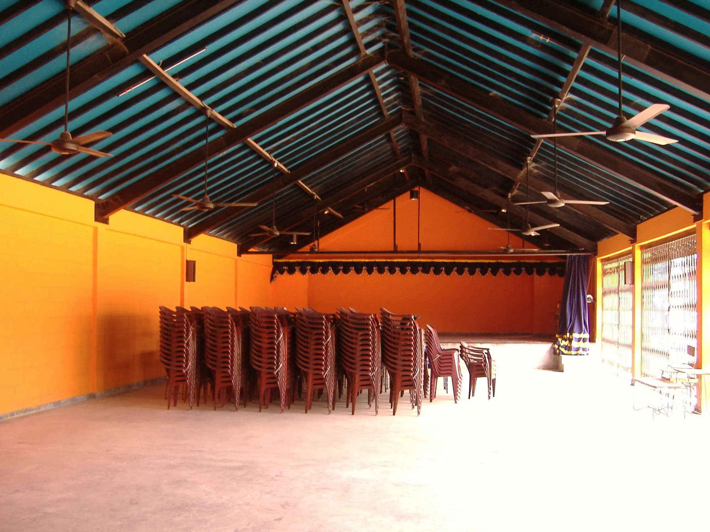 A hall under a pitched roof, with orange walls and stacked chairs.