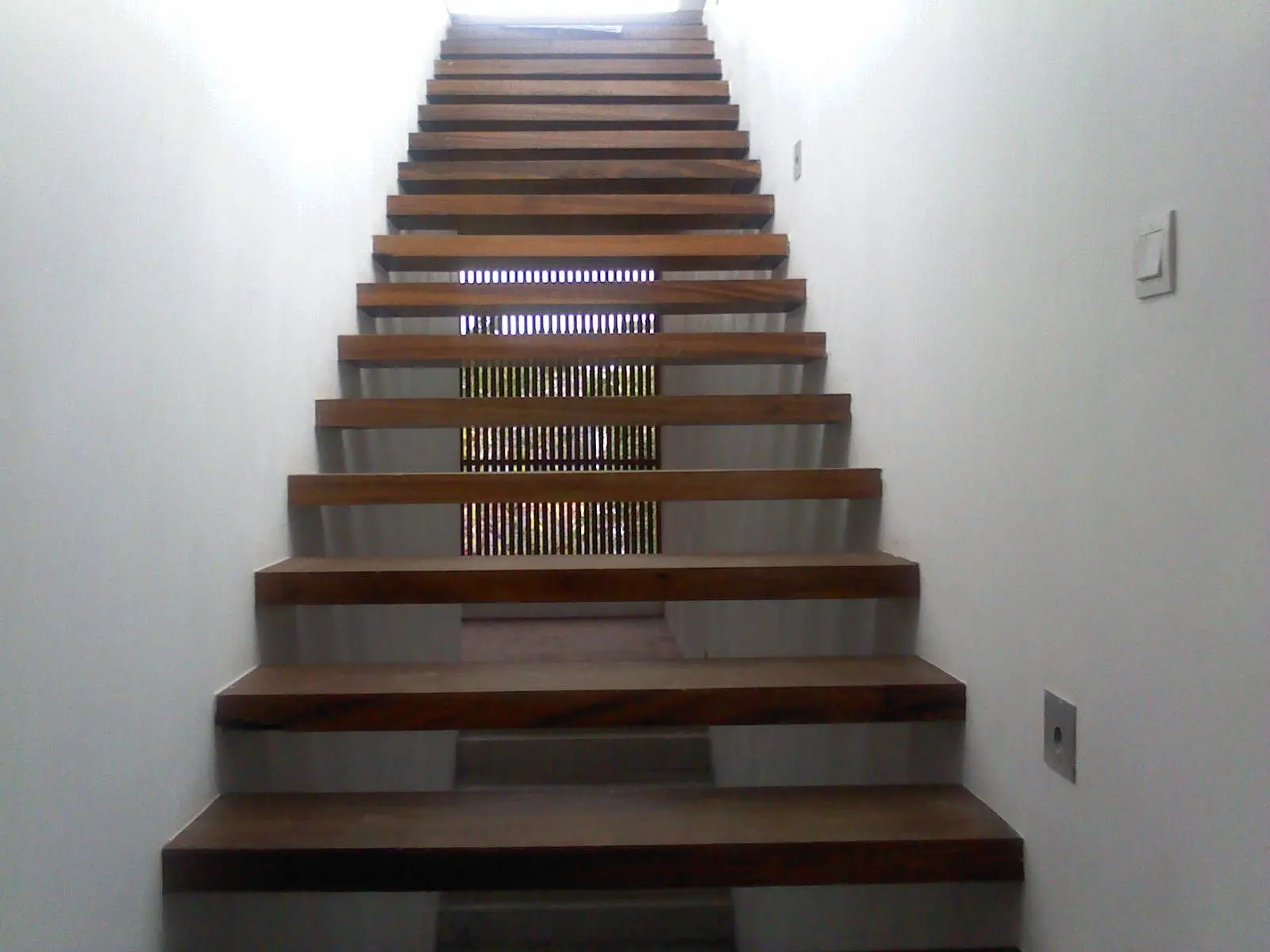 A straight stair beside a slatted screen in a white wall.