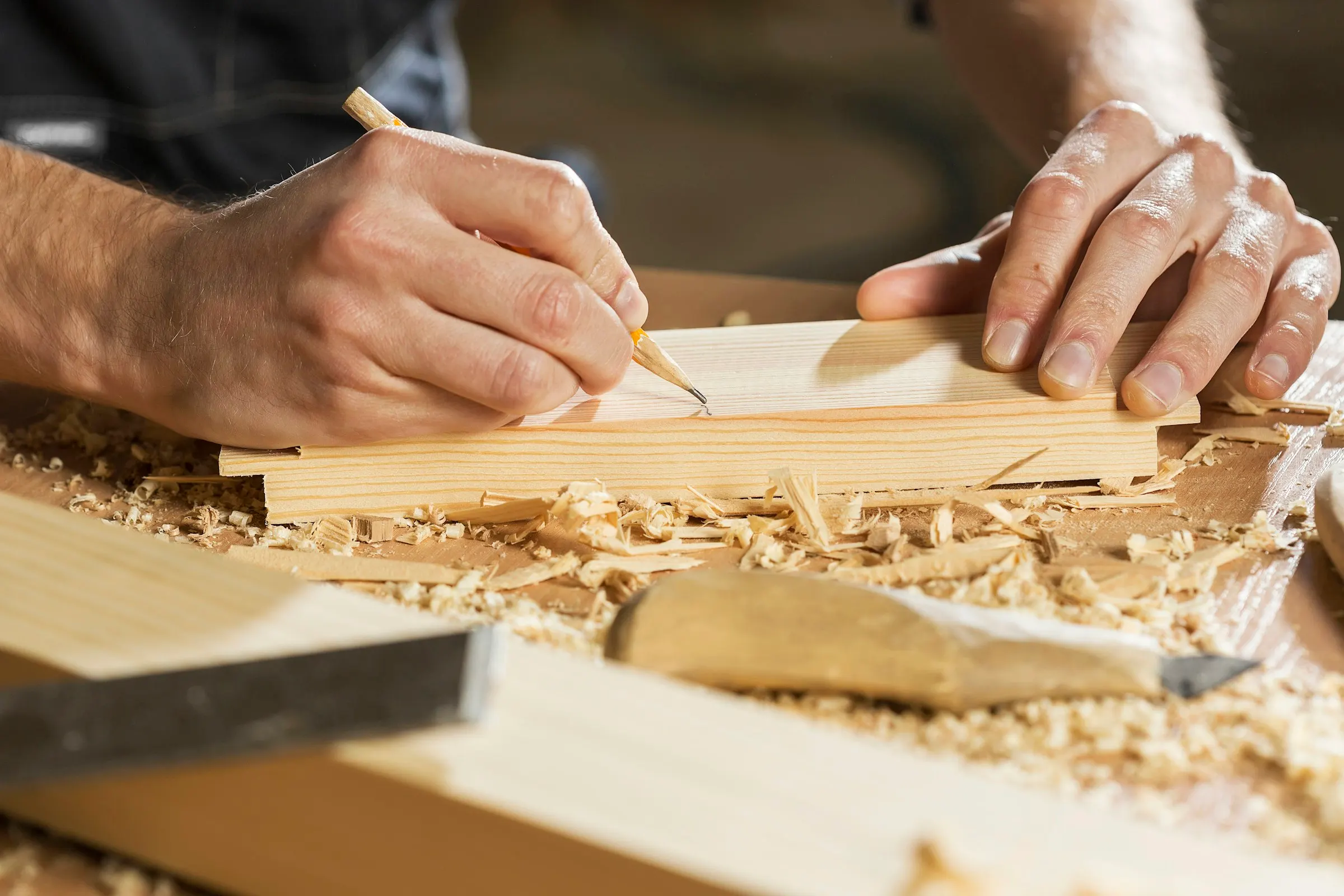 Hands marking a timber board with a pencil, wood shavings and a chisel on the bench.